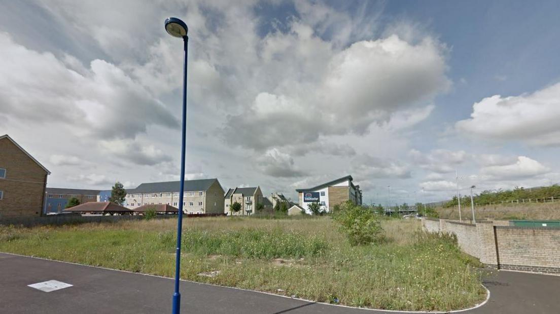 A piece of underdeveloped land with residential buildings behind and to its left side and a pavement with a blue street light in front