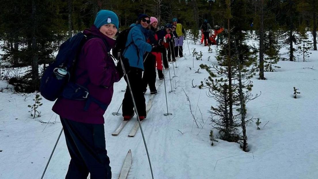 People in a line with ski equipment look back to face the camera. They are smiling, wearing hats and have bags on their backs.