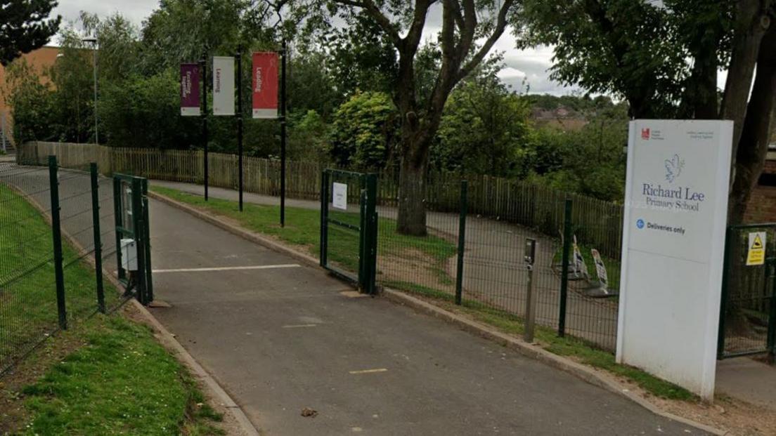 A tarmac path leads through a black gated fence, with grass areas on either side. There is a white sign before the gate that says "Richard Lee Primary School" in blue letters. 