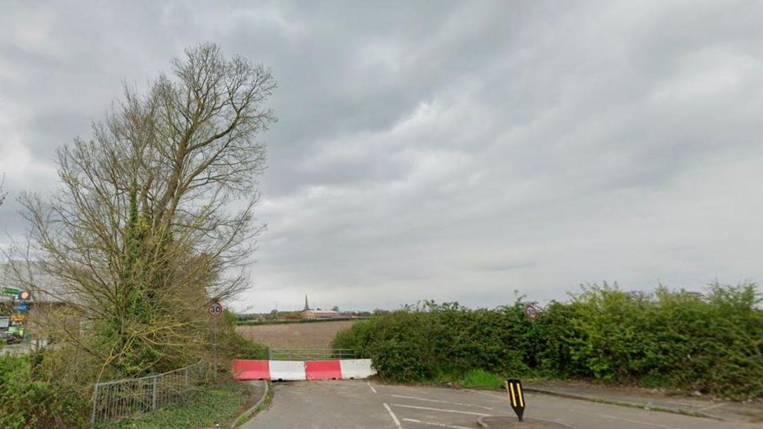 Field with bushes and trees around the border and a red and white barrier preventing access