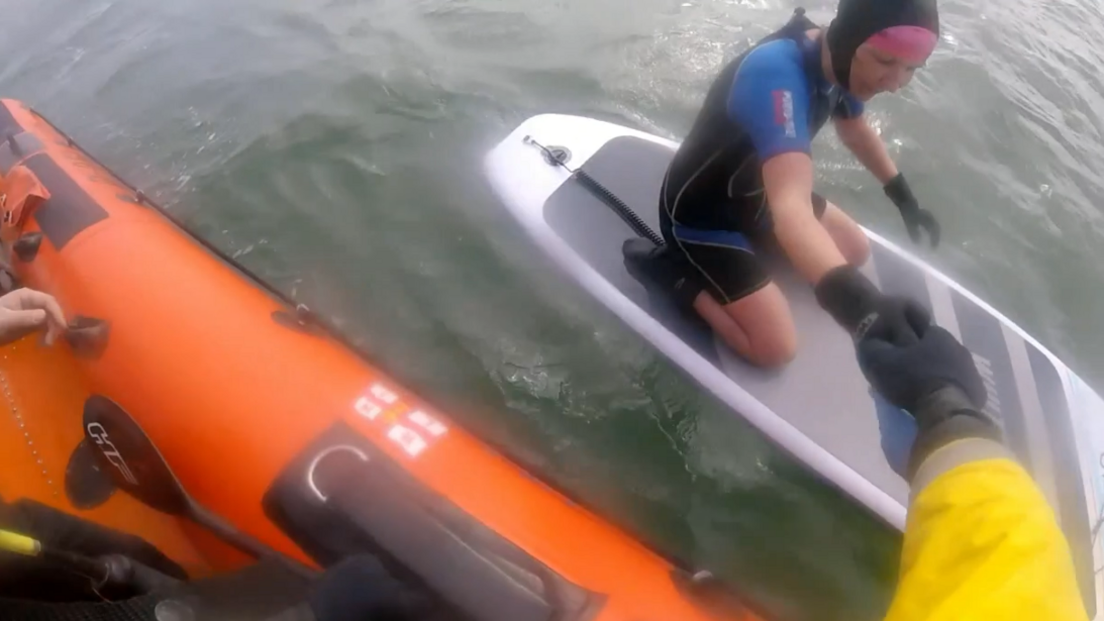 A woman in a wetsuit kneels on a paddle board in the sea, while a man in a yellow waterproof outfit pulls her onto an orange inflatable lifeboat.