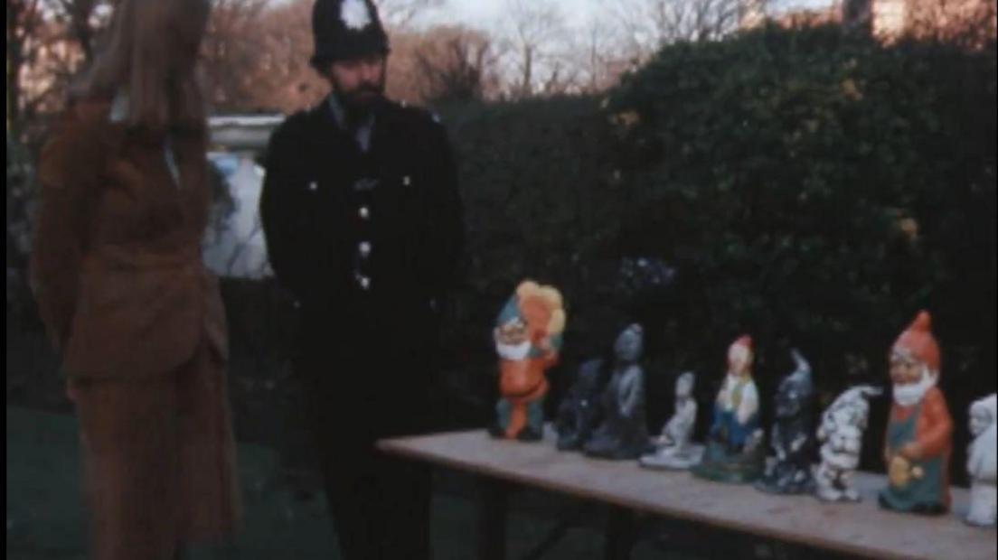 A policeman and a woman dressed in brown stand next to a wooden table in a garden. The returned gnomes stand on the table.