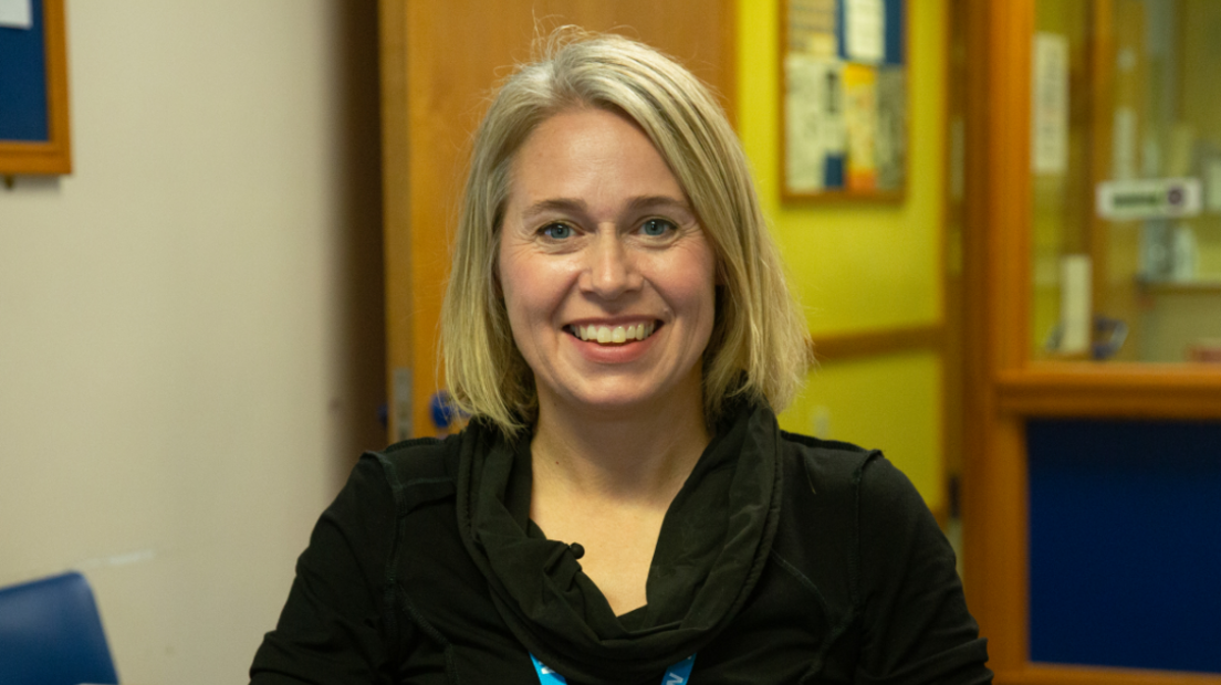 Dr Helen Munro is smiling and has a blonde bob-cut. She is wearing a black long-sleeve top. Sat in front of a clinical background and you can see a hint of a light blue NHS lanyard around her neck