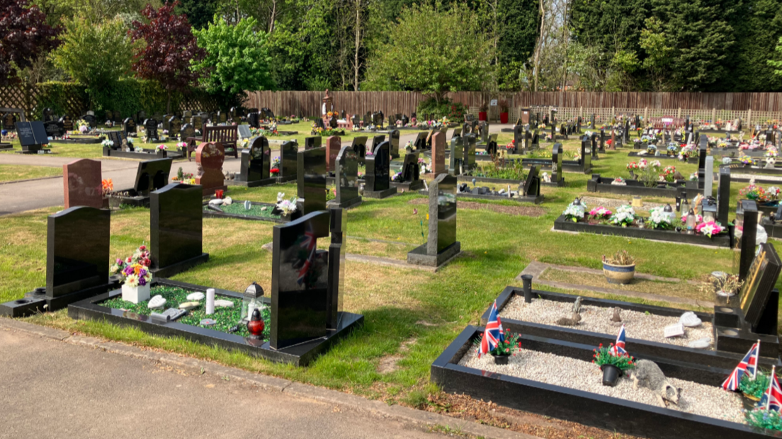 Rows of burial plots, most with black gravestones, set on a lawn, with a wooden fence and well-established trees in the background.