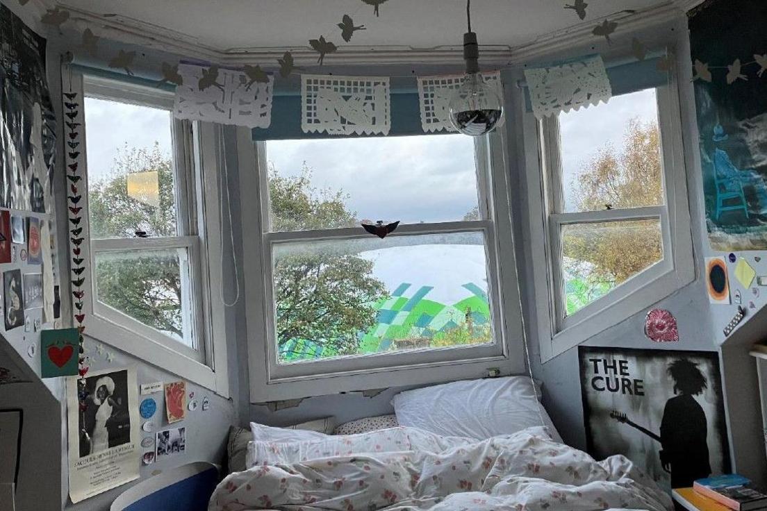 A bedroom covered in posters. There is a bay window and a bed underneath it. Most of the view through the window is obstructed by the giant inflatable tennis courts.