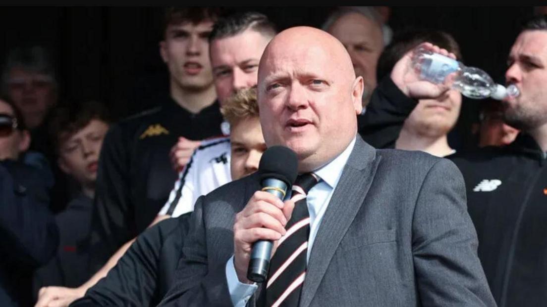A man with a bald head in a grey suit and black and white striped tie holding a microphone with other people behind him