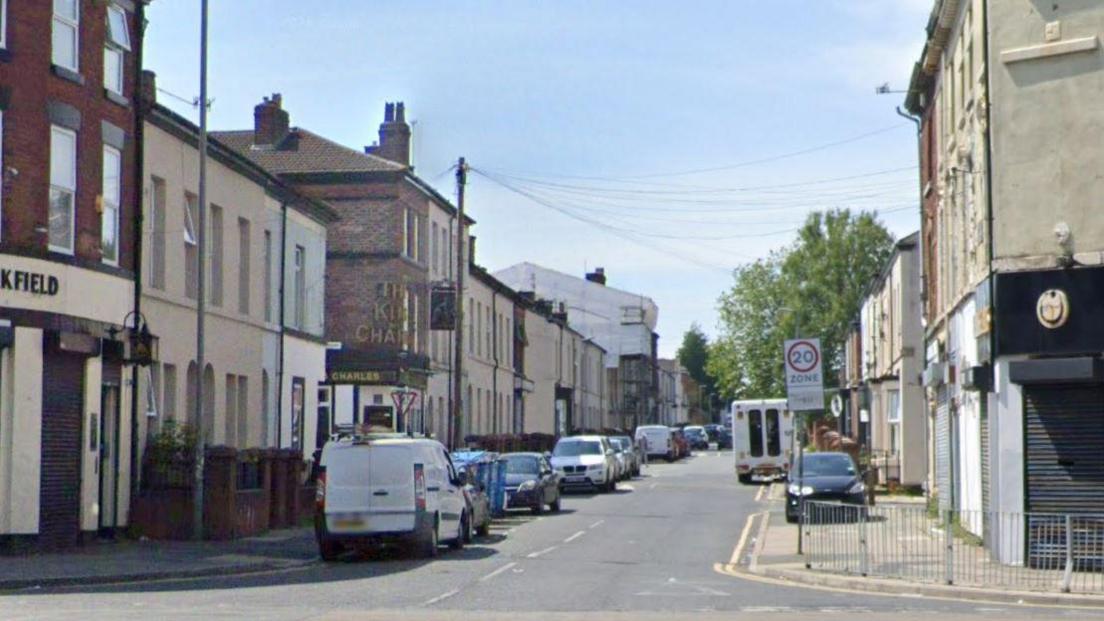 A road showing a mainly residential street with terraced houses and cars parked on both sides of the street.
