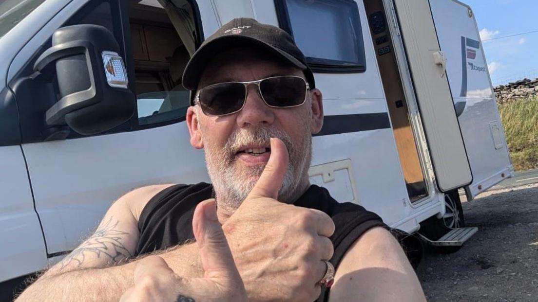 Bishop Mick Fleming wearing a black t-shirt, sunglasses and black cap holds his thumbs up. He is sitting in front of a motorhome parked near a grassed area and stone wall.