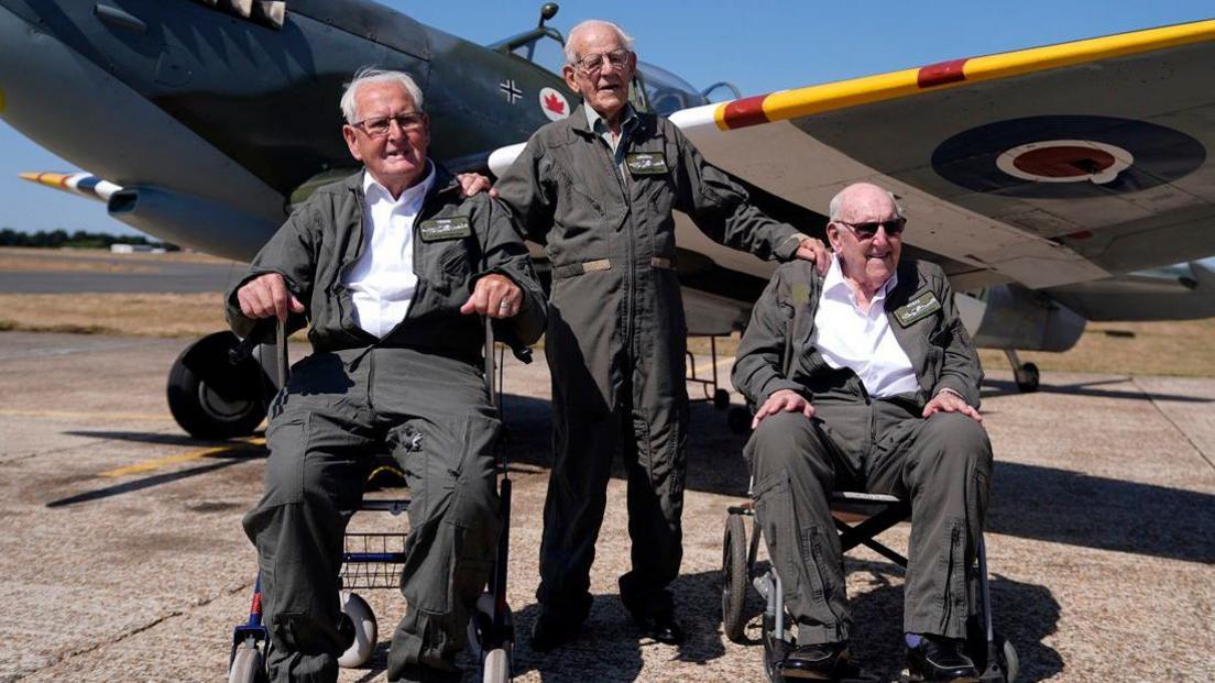 From left to right, Ivan Staines, 99, Paul Terry, 101, and Alfred Cooley, 100, pose in flying suits in front of a Spitfire at Biggin Hill Airport in Kent. Staines and Cooley are seated in wheelchairs while Terry stands between them with his hands resting on their shoulders. The aircraft is parked on the airfield behind them under a clear blue sky.