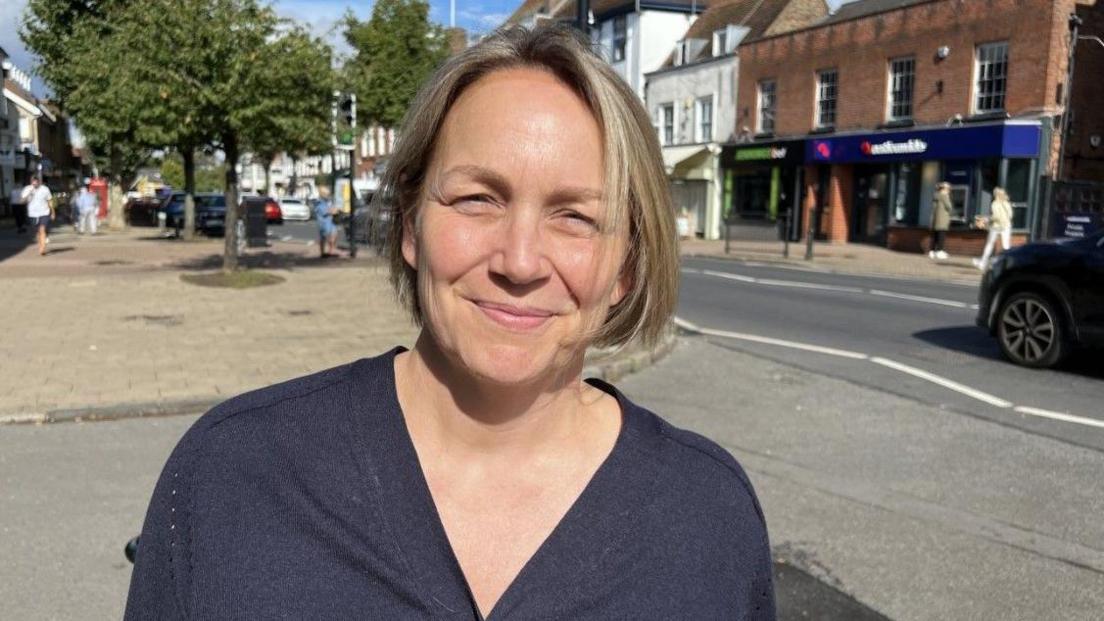 Kerry Gilroy is looking at the camera on a sunny day. She has a short blond bob and is wearing a dark blue top. Shops can be seen in the background as well as people walking on a street and a car on the road