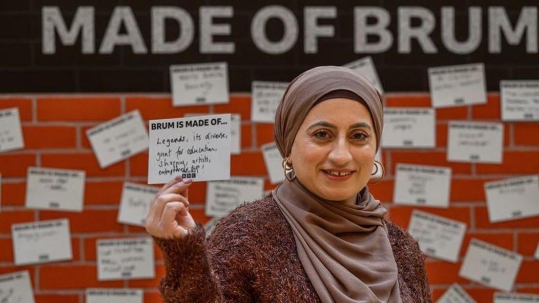 Fozia Fazil is looking at the camera. She is holding a piece of paper or card with words including Brum is made of legends, it's diverse. Other pieces of paper or card are on a red wall behind her.