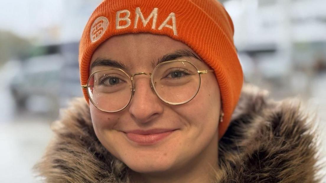 A young woman with an orange woolly hat which says BMA on it. She is wearing glasses and smiling into the camera. She has a coat with a furry hood