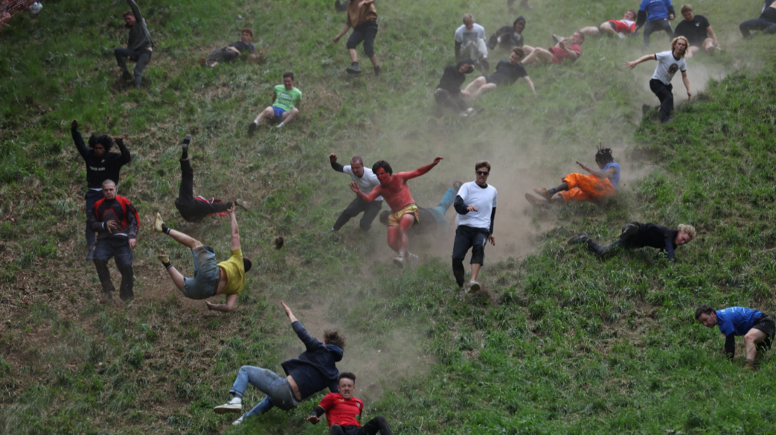 Image shows men running and tumbling down Cooper's Hill. The hill is very steep and grassy. The ground is so dry that there are dust trails behind the men as they run. Mot of the men have fallen over and are rolling and falling down the hill, with only a few of them still on their feet.