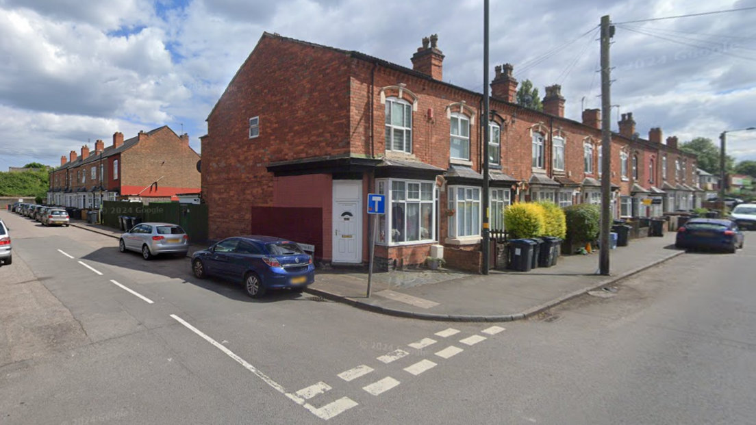 The junction of two roads, with a row of terraced houses stretching away from the camera, and cars parked alongside the side of the end terrace 