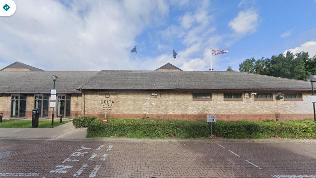 Single-storey brick-built hotel with small brown-framed windows and a grey pitched roof. There are entrance doors set back on the left. There is a low hedge in front of the building and a roadway in the foreground of the picture. There are trees in the background to the right.