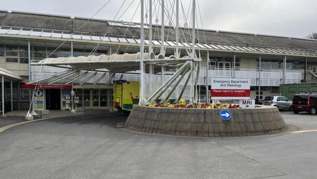 A white sign saying "Emergency Department and Radiology". A smaller white sign underneath which says "MRI". There is a small roundabout outside of the  entrance of the building. There is a yellow and green ambulance outside of the doors. 