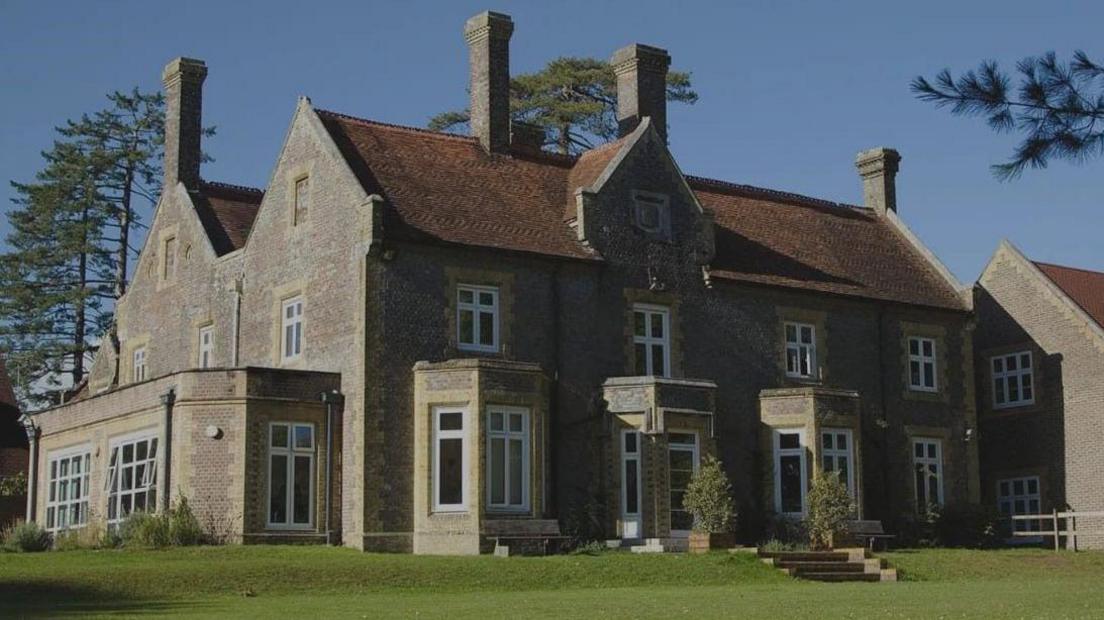 A grand old building that's now used as as a school with 6 chimneys and steps leading down to a grass lawn 