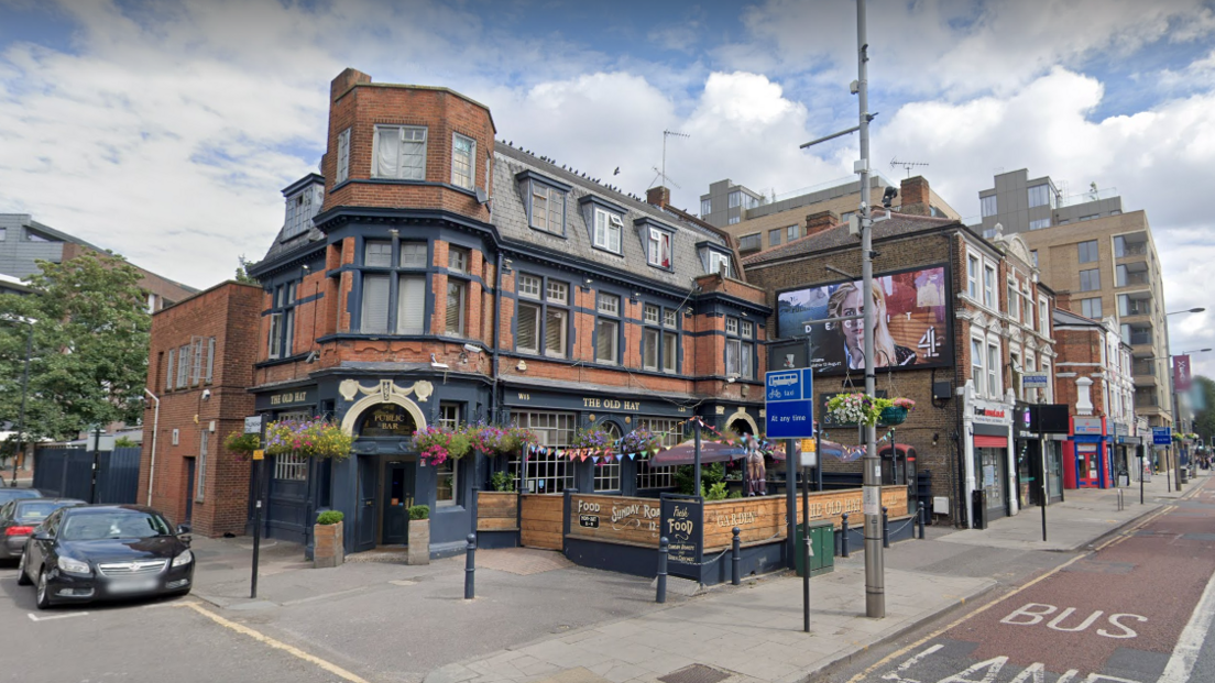 Street view of the outside of The Old Hat pub in Ealing, a brick building with a turret, dormer roof and hangings baskets of flowers, with a dark painted frontage and window frames, and a fenced off outdoor area 