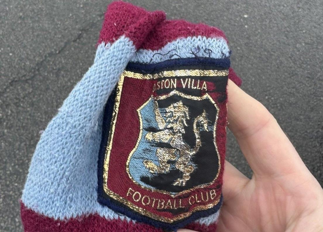 A close-up of the claret and blue-striped scarf, held in a hand. The scarf features an embroidered club crest of a golden lion on a claret background. Surrounding the lion are the words Aston Villa Football Club. The scarf is held in a hand, with the picture ending at the wrist.