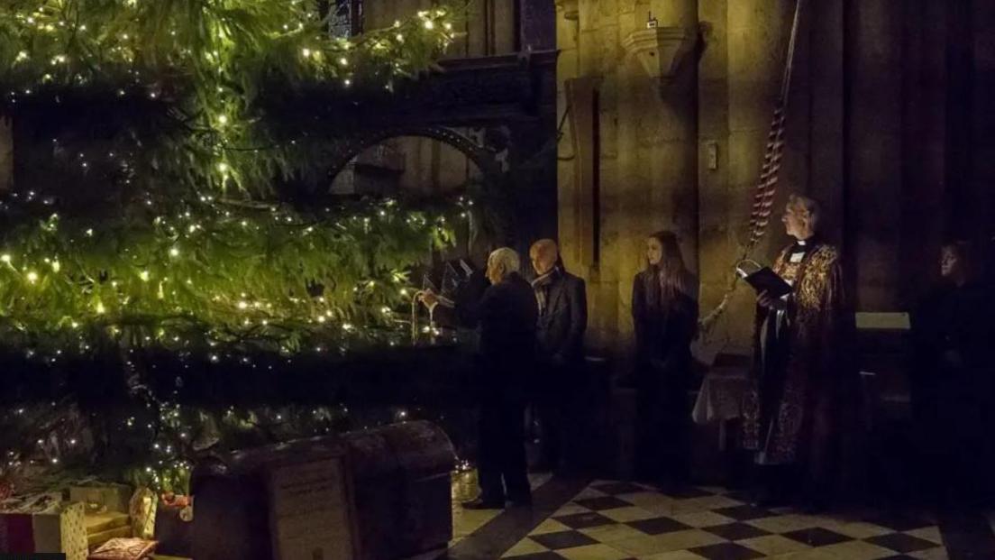 Jackie McCowliff hanging the miner's safety lamp on the tree. He has white hair. Among those also present are Reverend Canon.
