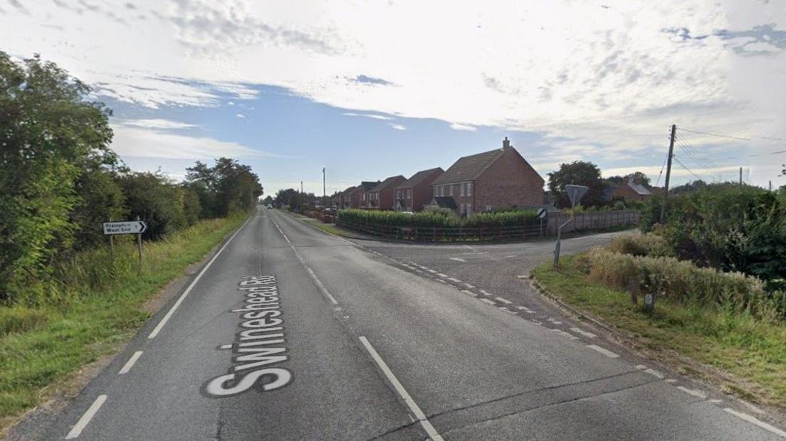 A Google Images screengrab of the junction where the collision happened, it is an A road with signs pointing towards two villages to the left hand side of the road. There are hedges and houses.