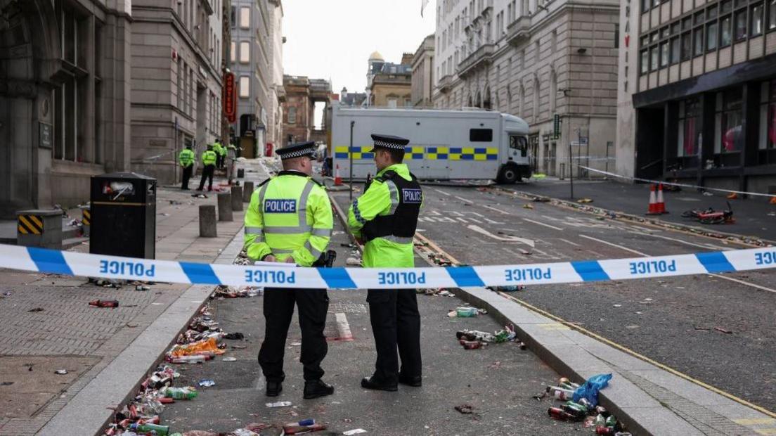 A police cordon is in place on Water Street. Two police officers are standing behind the cordon and there are a number of officers located further down the road. A police van is parked across the road.