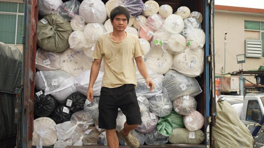 A man in a beige t-shirt and black shorts is stepping down from a truck piled high with rolls of fabric of various colours - pink, white, green. 