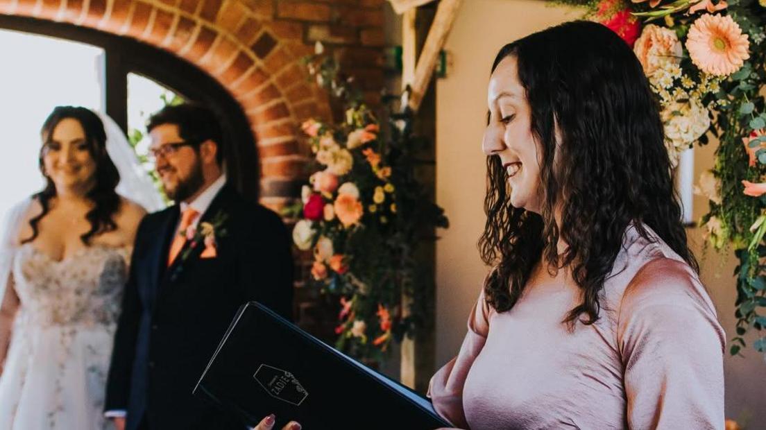 Zadie Ward, in a pink dress, reads from a black folder at a wedding