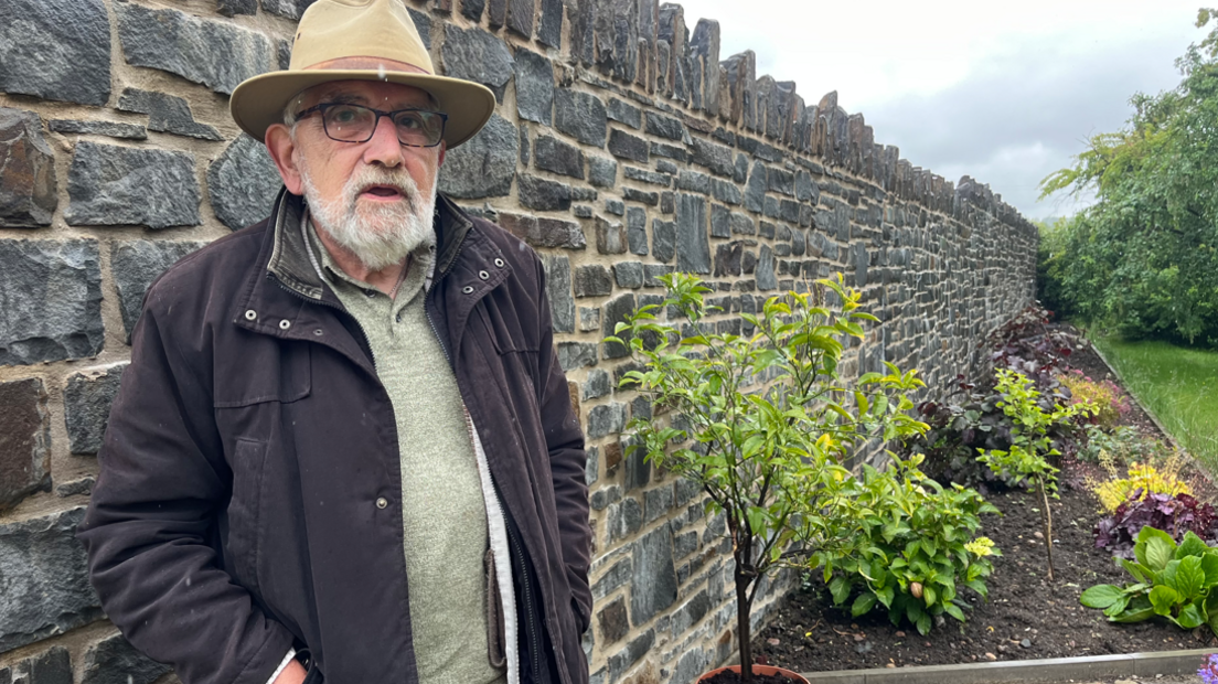 John Watkeys standing against his new stone garden wall which is about head height. He is wearing a fawn-coloured hat and glasses with a black coat and has a white beard. A flower bed can be seen to the right of the wall and grass to the right of that.