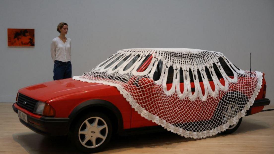 A gallery assistant poses with Turner Prize shortlisted artist Jasleen Kaur's installation, a doily covered red Ford Escort