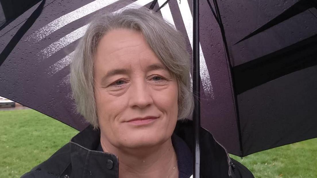 The picture shows the Lib Dem MP Claire Young for Thornbury and Yate standing under an umbrella on Cranmoor Green in Pilning. She has short grey hair and is wearing a dark-coloured jacket