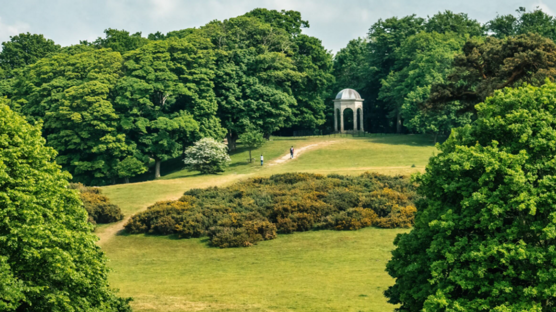 A green park with two people on a path walking uphill to a folly. The open area is surrounded by trees. 