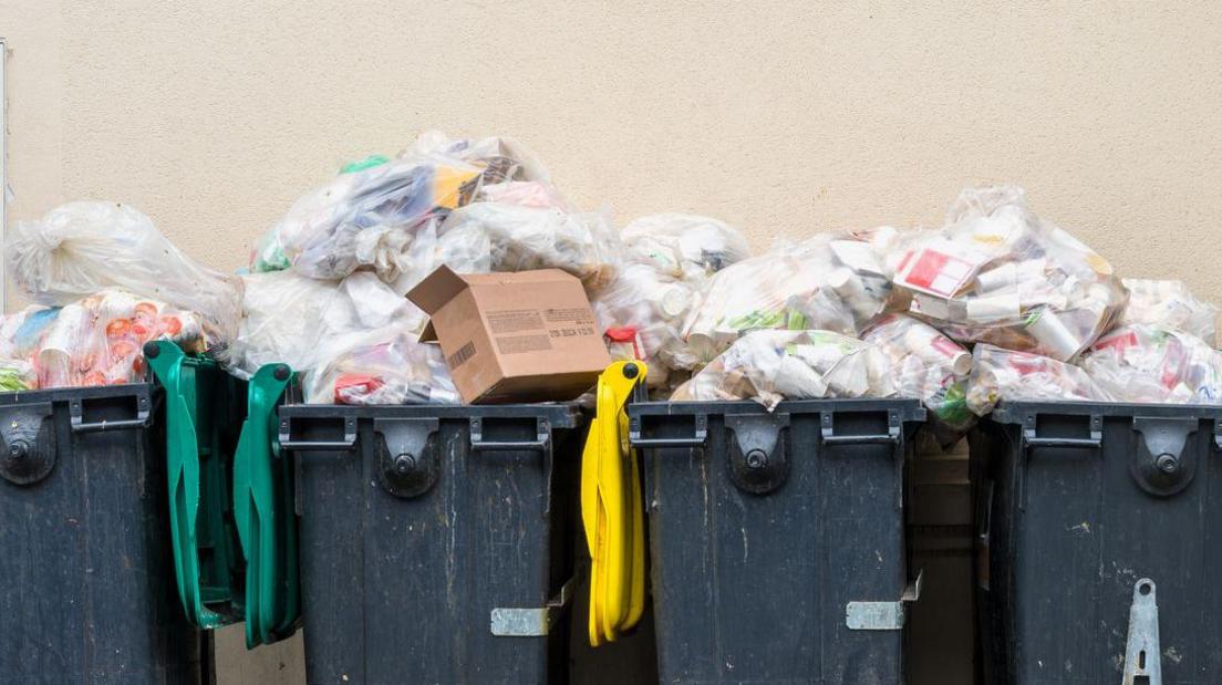 Four large grey bins outside against a beige wall. They are overflowing with rubbish.