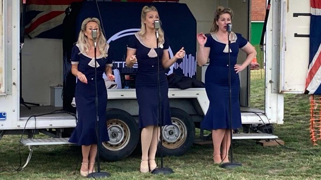 Three women wearing blue dresses. They are standing on grass in front of microphones.