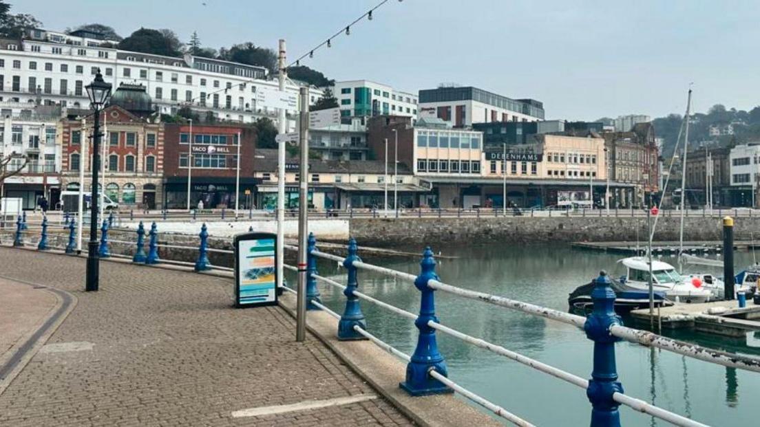 Torquay harbourside in March 2025. There is a row of buildings overlooking the water and a pavement to the left, with a white and blue barrier along the water's edge.