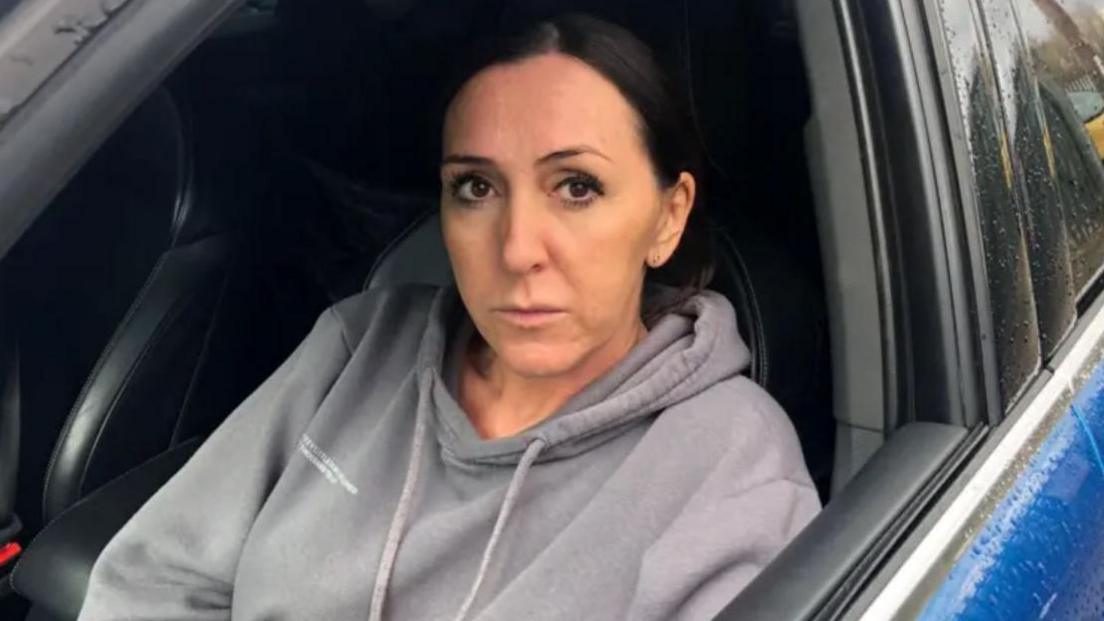 A woman in a grey hoodie with black hair, pictured in the driver seat of her car through the open driver's side window. Her car is blue.