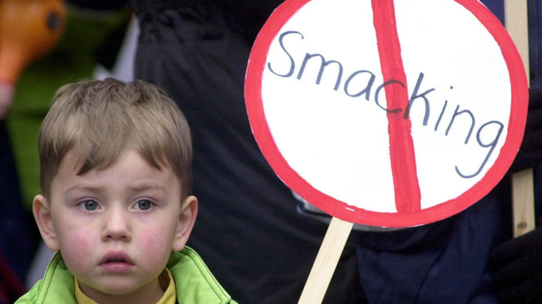 A young child holds a placard with the word smacking crossed out