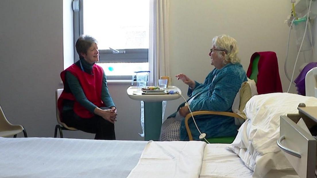 Volunteer Isabel sits with a patient in Newton Abbot Community Hospital. Isabel is wearing a red tabard. She is speaking to a female patient in a blue dressing gown. 