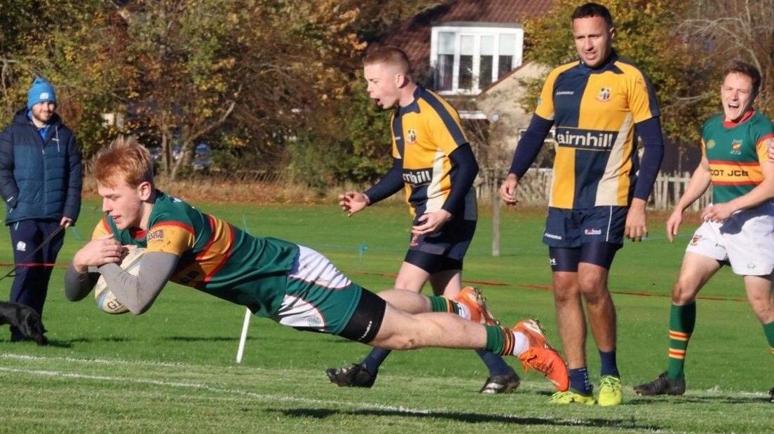 A man is in mid air as he scores a rugby touchdown. He has ginger hair and is wearing a green and yellow strip.