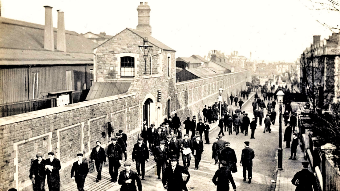 A black and white photo of a street lined by houses on the right and a long stone wall with an archway on the left. Behind the wall are large buildings, some with tall chimneys. Many men in dark coats and flatcaps are walking in the street.