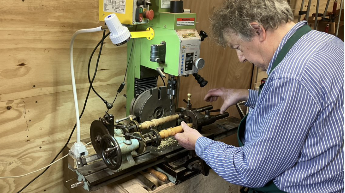 Daniel Bangham, who has grey hair and is wearing a blue and white striped long-sleeved shirt, stands at a drilling machine. He holds a piece of woodwind instrument in place while it is held at both ends by a metal clamp. A drill, held by the machine, hovers above the wood.