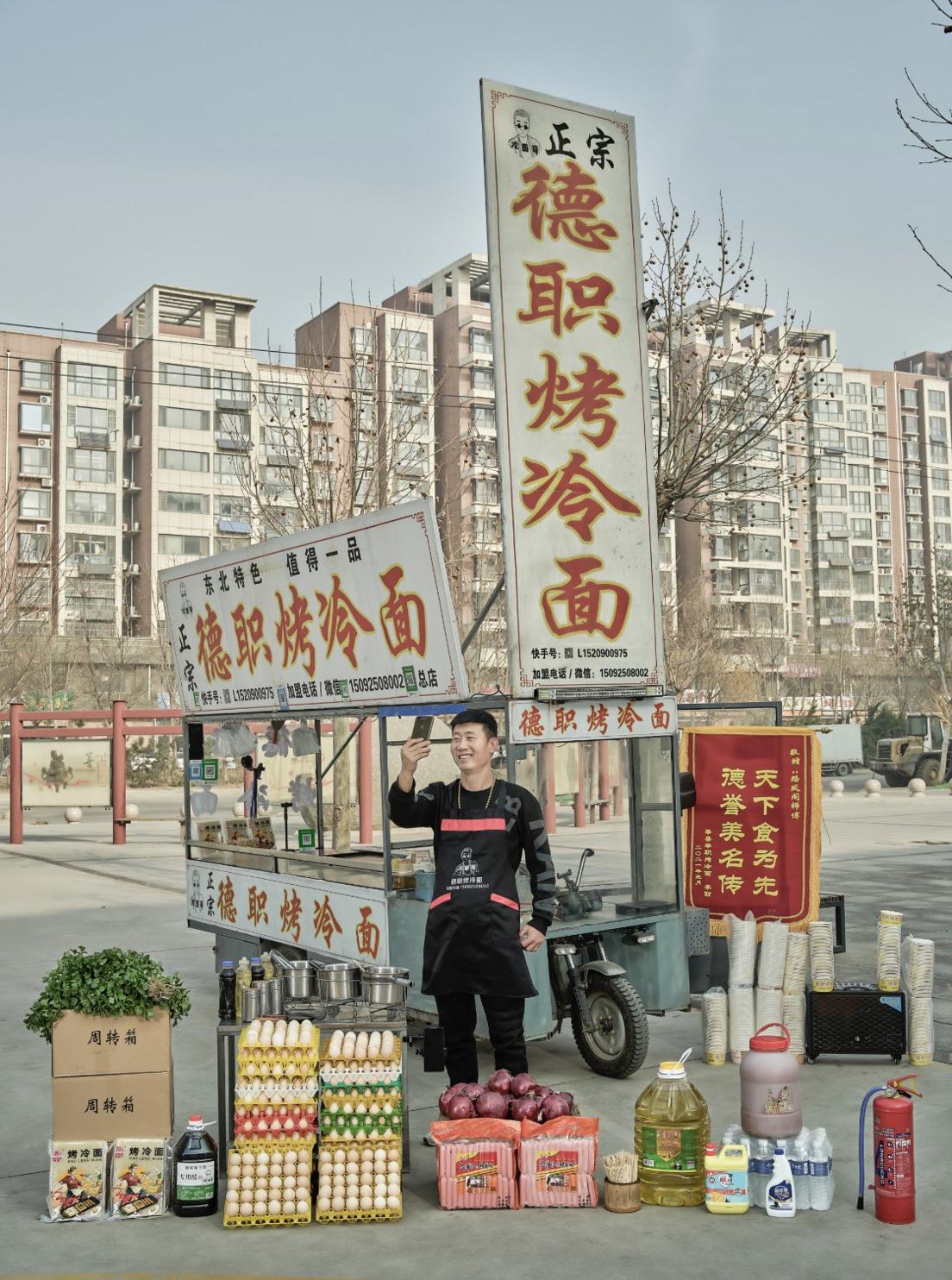 Man stands in front of a roadside hawker stall while taking a shot of himself. In front of him are trays of eggs, cooking oil, bottles of sauces and produce to cook cold grilled noodles.