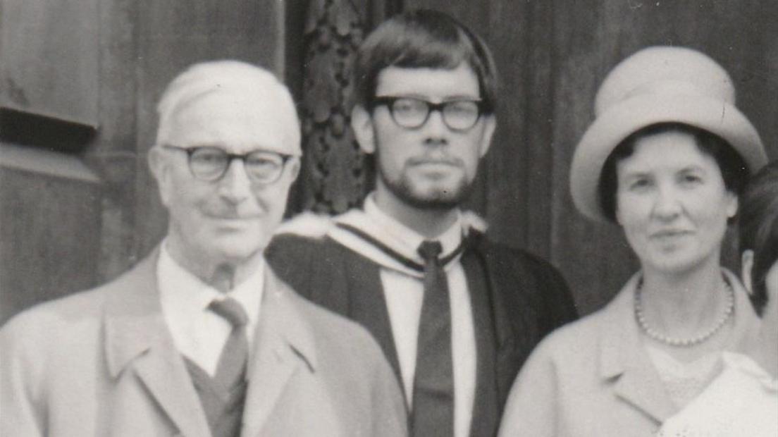 A black and white photograph of John Harrison pictured in 1965. He is dressed in a suit and graduate gown and has dark hair and dark framed glasses. A man and a woman are stood either side of him. The man is wearing a suit and coat and dark framed glasses. The woman is wearing a light coloured coat and hat.