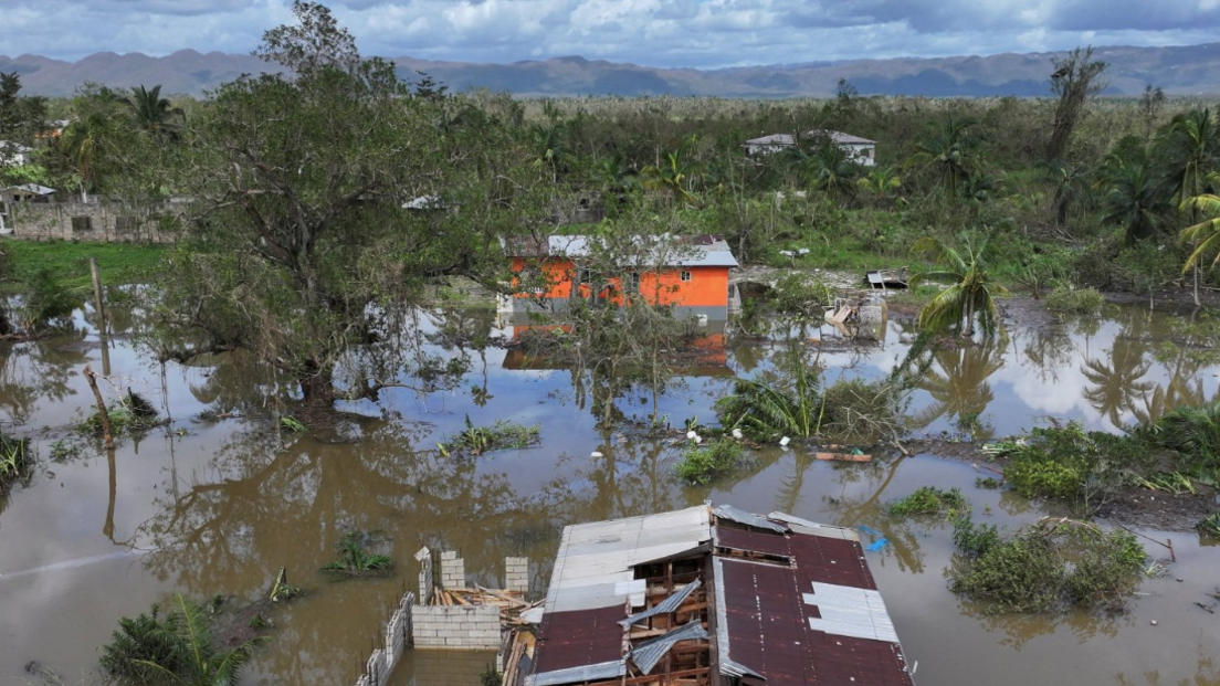 A desolate scene showing the destroyed roof of a building surrounded by water, with fallen trees scattered around. 