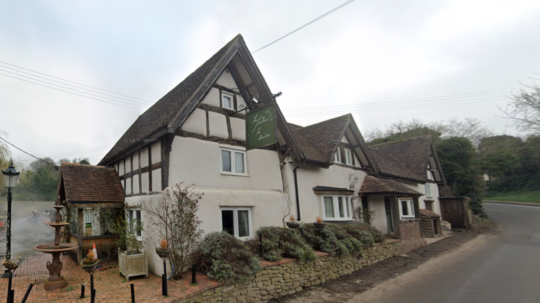 A white pub with brown tiled pointed roofs site beside a village road. There is a green sign hanging from the building that says 'The Feathers Inn in Brockton". There are bushes and a small brick wall in front of the building.