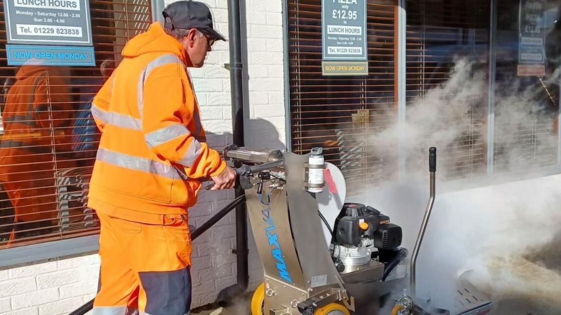 A council worker in a cap and high-vis orange overalls is pushing a street cleaning machine. There is smoke rising from the ground. 