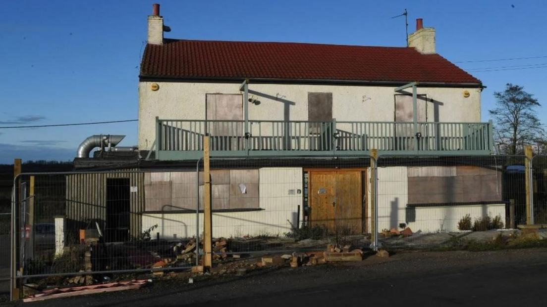 The exterior of the Dog in a Doublet pub after it closed. It is a white painted two-storey brick building, with a green painted balcony. Its windows and doors are boarded up. There is fencing around it.