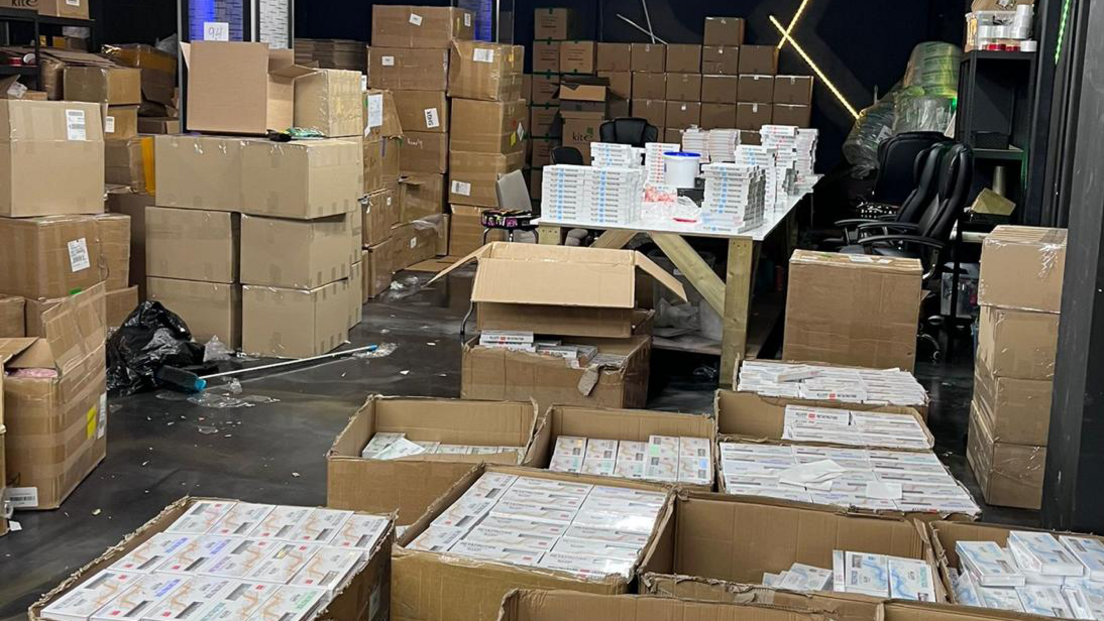 Many brown boxes, piled up in a warehouse, full of weight loss packaging. There is a table, chair and shelves in the distance.