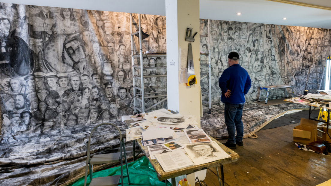 A man stands in front of a wall with his hands on his hips. On the wall is a large swathe of fabric, covered in hundreds of faces drawn in charcoal. In the foreground there is a table with art material on, and a small stepladder.