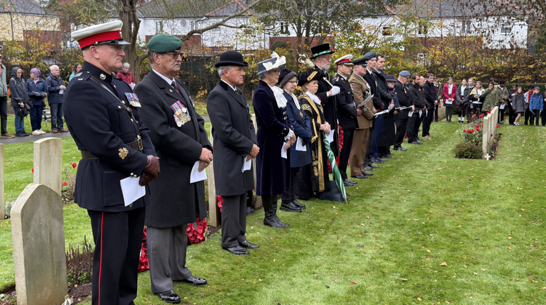 Veterans and members of the local authority are gathered in a cemetery all wearing their uniforms.